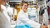 Woman in a white coat smiling in an industrial laboratory in front of machines.