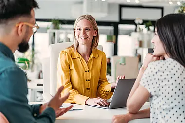 Eine Frau sitzt mit zwei weiteren Personen am Tisch und unterhalten sich gemütlich.